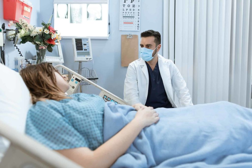 a doctor sitting down in a white lab coat and a mask and talking to a patient on a bed